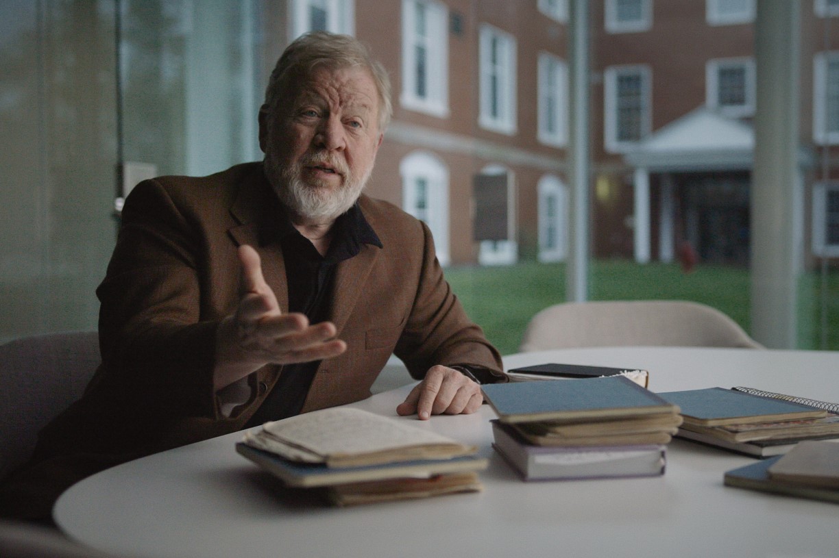 Dan Everett, in a black shirt and brown blazer, sits at a table surrounded by notebooks.