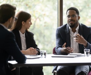 Photo of Black male executive at head of conference room table, talking and gesturing to two colleagues.