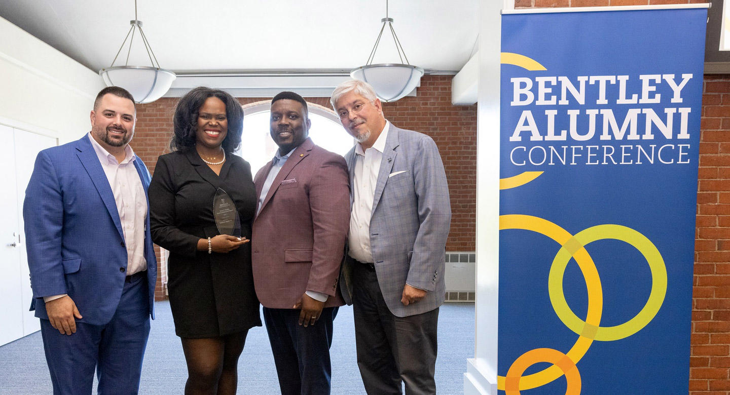 four alumni pose next to the inaugural Bentley Alumni Conference banner