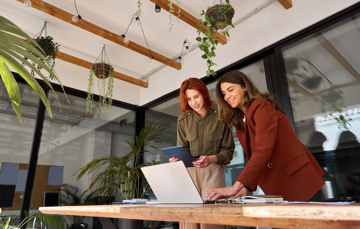 Two happy business women of young and middle age talking working in green office. Smiling professional ladies entrepreneurs partners using laptop and tablet technology devices standing at work table.