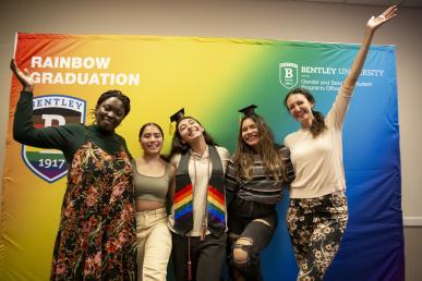 Bentley students and staff celebrate Rainbow graduation in front of rainbow backdrop