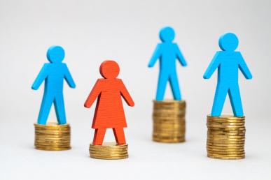 Photo illustration of four wooden figures — three male and one female — balance atop individual stacks of coins, with the female figure's stack noticeably smaller than all three of the men's