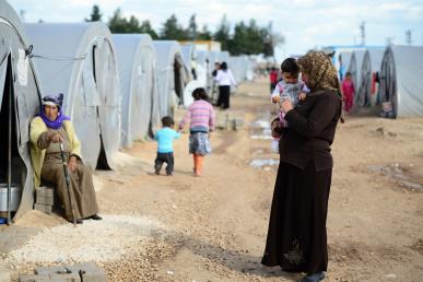 A Syrian woman wearing a kaftan and headscarf holds an infant as she stands in the middle of dirt pathway of a tented refugee camp.